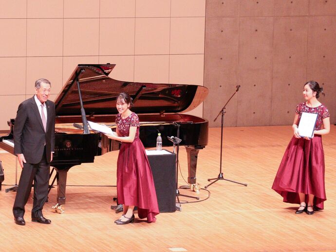 なぎさホールの舞台の上に市長と山田姉妹。山田姉妹は市長から手渡された委嘱状を持っている。