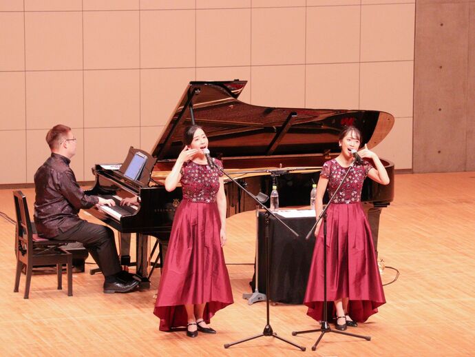 なぎさホールの舞台の上にグランドピアノ。ピアノの前には山田姉妹が立っており、二人で手話を交えて歌っている様子。